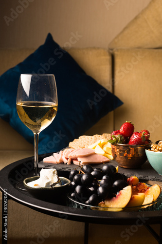 Cheese and fruits plate with glass of white wine on round tray, colorful and delicious charcuterie with snacks and wineglass for romantic evening, wine tasting, selective focus.