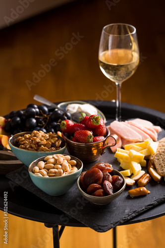 Cheese and fruits plate with glass of white wine on round tray, colorful and delicious charcuterie with snacks and wineglass for romantic evening, wine tasting, selective focus.