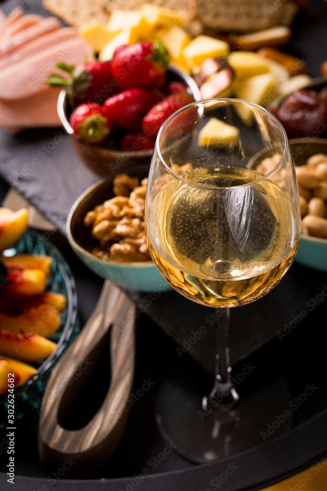 Cheese and fruits plate with glass of white wine on round tray, colorful and delicious charcuterie with snacks and wineglass for romantic evening, wine tasting, selective focus.