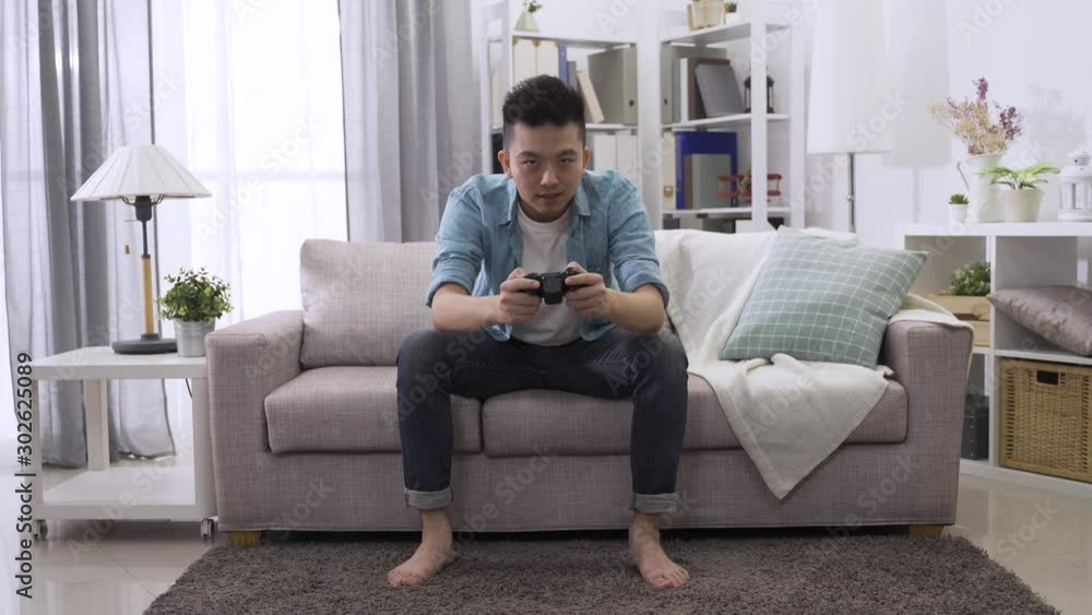 Man in casual wear sitting on sofa and playing video game in home living room. asian korean guy addicted to tv entertainment. lazy male holding joystick control having fun laughing on couch indoors. Man in casual wear sitting on sofa and playing video game in home living room. asian korean guy addicted to tv entertainment. lazy male holding joystick control having fun laughing on couch indoors.