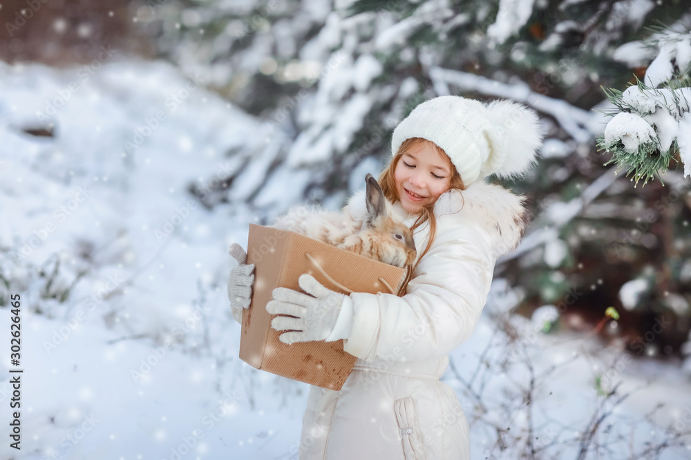 A girl in a white fluffy down jacket in a snowy forest holds a decorative live rabbit in a box. A girl in a white fluffy down jacket in a snowy forest holds a decorative live rabbit in a box.