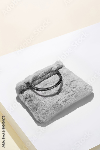 Object shot of a grey faux fur bag with metallic semicircular bag handles, lying on a white surface. The bag is casting a shadow.