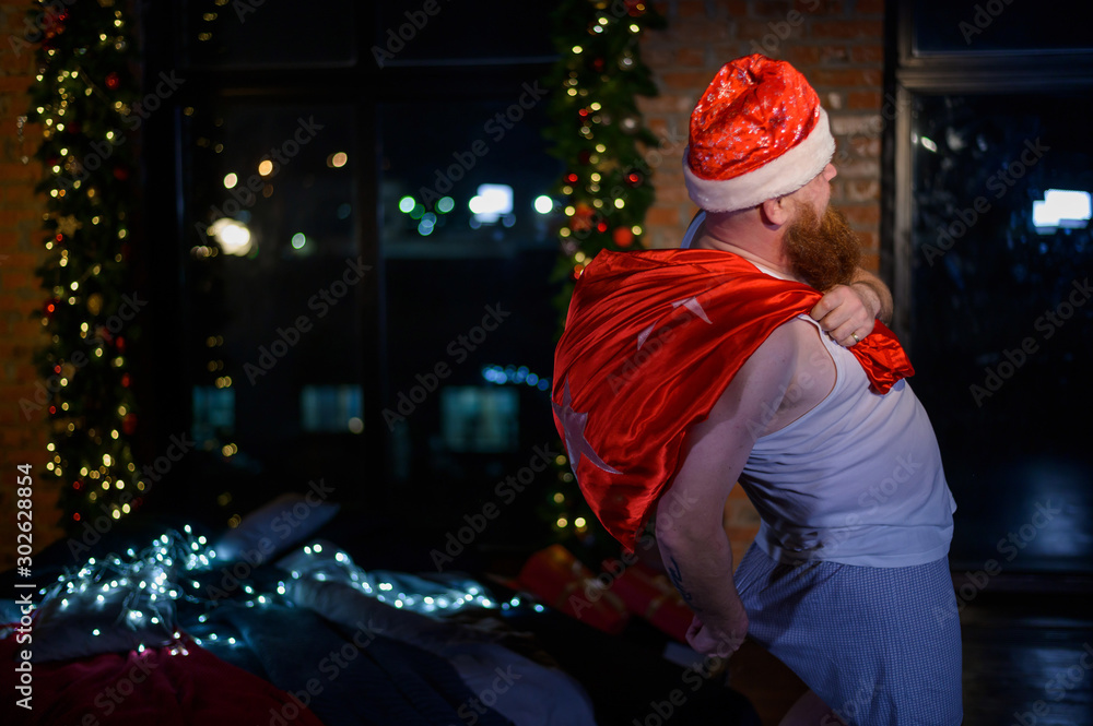 Bad angry santa with a red beard carries a bag with gifts on Christmas Eve. A man in shorts and a santa hat scratches his butt. Santa Claus before the New Year. Bad angry santa with a red beard carries a bag with gifts on Christmas Eve. A man in shorts and a santa hat scratches his butt. Santa Claus before the New Year.