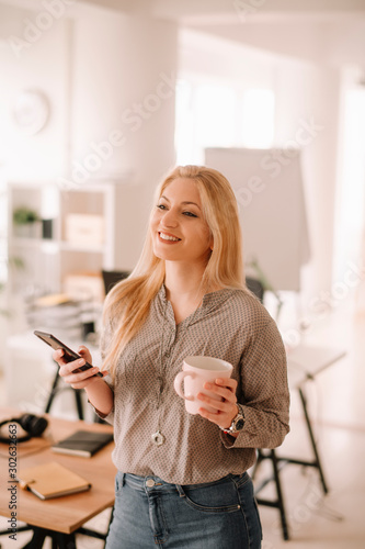 Young woman in office. Beautiful woman on coffee break using phone. 