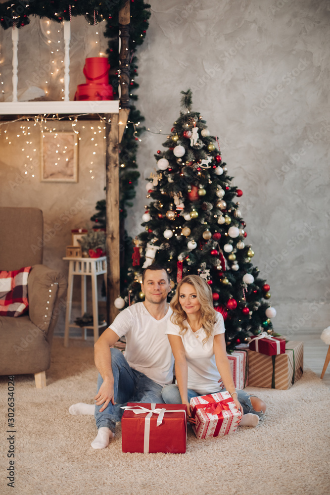 Stock photo portrait of beautiful young couple in similar clothes sitting on cozy beige carpet with wrapped Christmas presents and smiling at camera. They are sitting on the floor against beautiful Stock photo portrait of beautiful young couple in similar clothes sitting on cozy beige carpet with wrapped Christmas presents and smiling at camera. They are sitting on the floor against beautiful