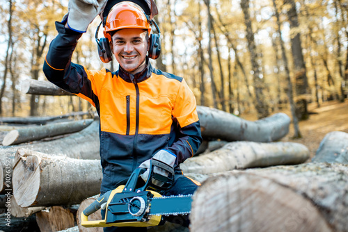 Portrait of a cheerful professional lumberjack in protective workwear standing with a chainsaw on a pile of logs in the forest photo