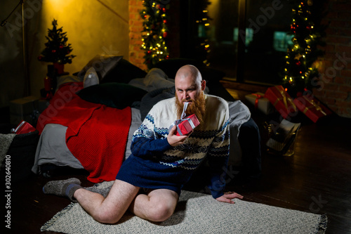 Humorous image of a man in a sweater and without pants in front of Christmas. Bald with a red beard unpacks a gift. A parody of a female photo shoot. Joke.