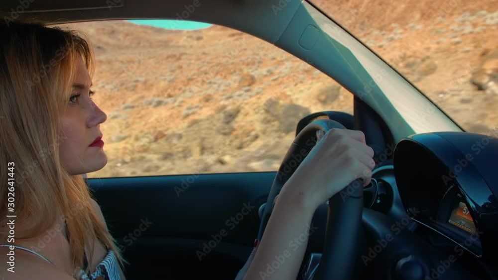 Attractive blonde woman drives a car, holds the steering wheel. Outside the window is a desert landscape of a volcanic island. Concept woman drives a car, gender equality. Attractive blonde woman drives a car, holds the steering wheel. Outside the window is a desert landscape of a volcanic island. Concept woman drives a car, gender equality.