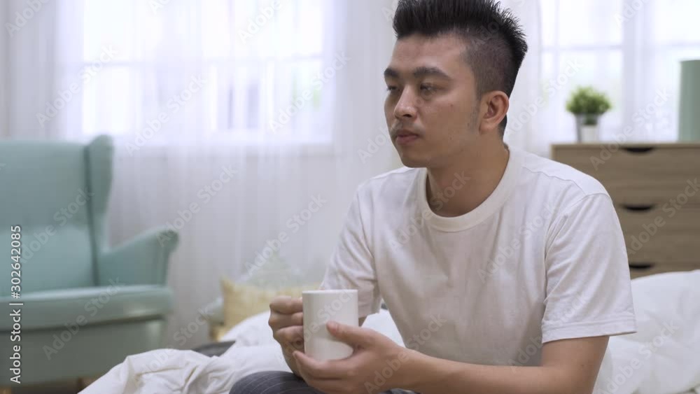 Young asian chinese man sitting in bed drinking water in mug. male enjoy breakfast beverage in early morning bedroom. attractive guy in tshirt relax and having cup of coffee to keep him awake at home Young asian chinese man sitting in bed drinking water in mug. male enjoy breakfast beverage in early morning bedroom. attractive guy in tshirt relax and having cup of coffee to keep him awake at home