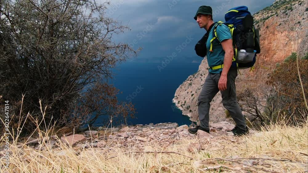 A man caucasian tourist with a backpack walks along a mountain trail against the background of the sea and cloudy sky during a hiking trip. Rest at nature A man caucasian tourist with a backpack walks along a mountain trail against the background of the sea and cloudy sky during a hiking trip. Rest at nature