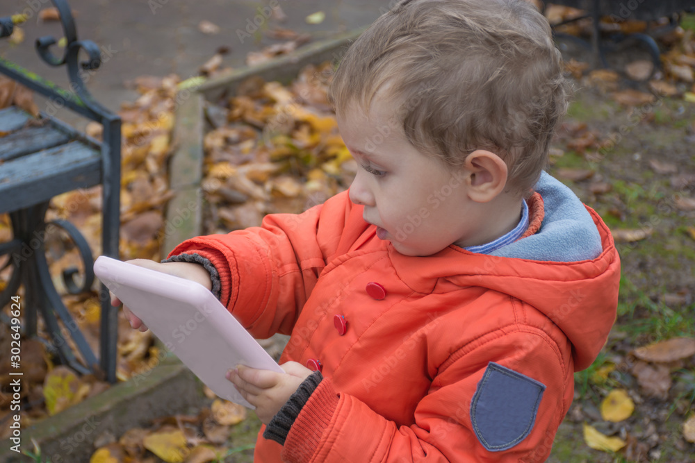 Little caucasian boy, 3 years old, in orange jacket and jeans is going to school. Child used laptop on park. Kid play game on laptop. Preschool education knowledge concept. Little caucasian boy, 3 years old, in orange jacket and jeans is going to school. Child used laptop on park. Kid play game on laptop. Preschool education knowledge concept.