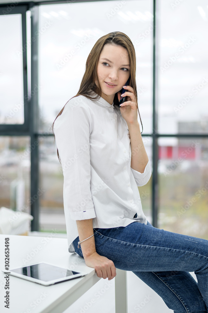 A young brunette in a white shirt and jeans writes SMS on the phone. The  girl