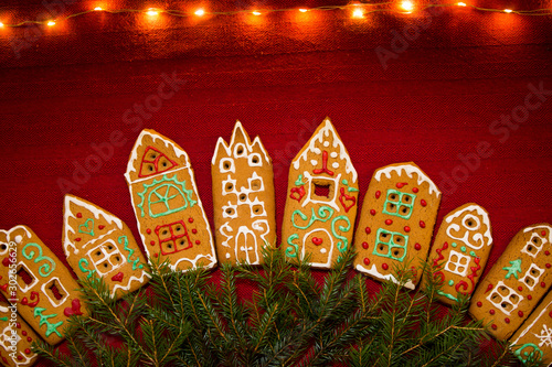 Christmas background. Red table cloth with town of cute gingerbread houses decorated with icing, Christmas lights, glitter and fir tree branches. Holiday mood. 