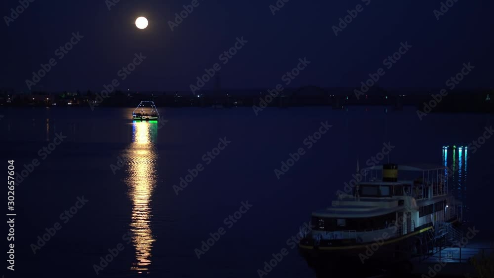The ship floats on the river at night. The moon is shining. On the water the lunar path. Ship glow with colored lights The ship floats on the river at night. The moon is shining. On the water the lunar path. Ship glow with colored lights
