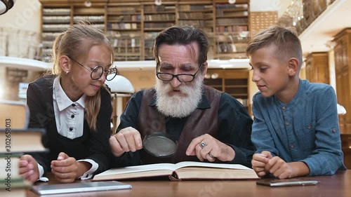 Intelligent eldery man with well-groomed beard sitting at the library table and reading book with his teen likable grandchildren using magnifying glass