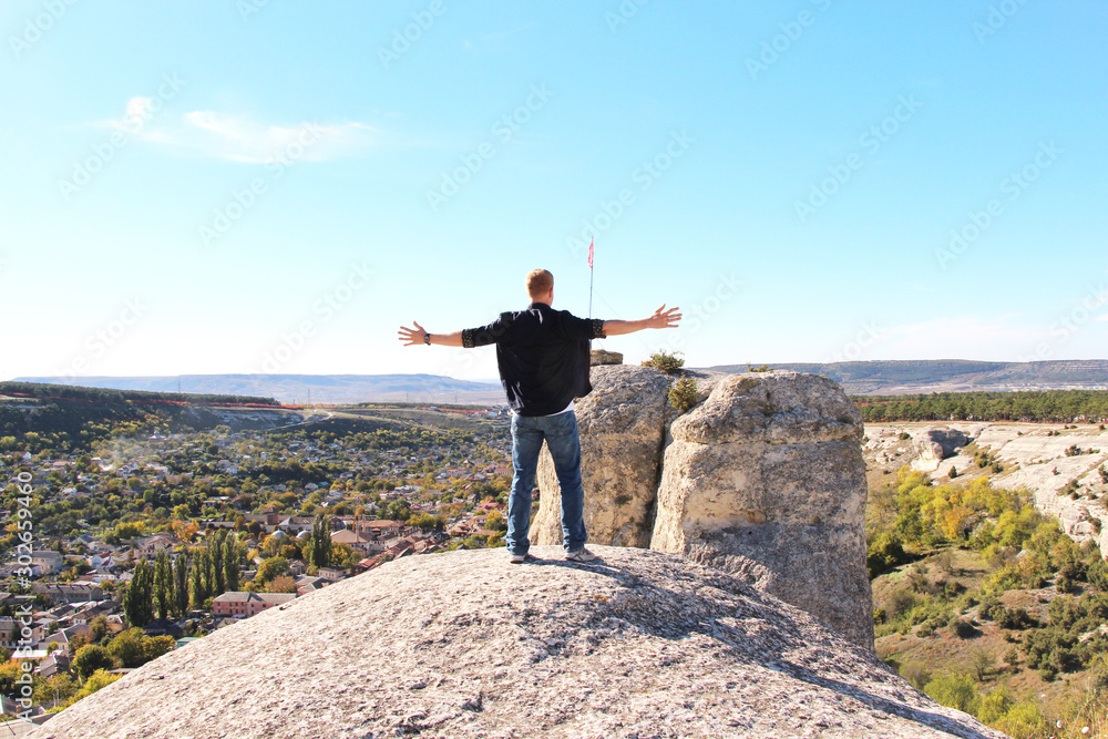 Obraz premium Redhead man stands with open arms on the top of Bakhchisarai Stone Sphinxes above Bakhchisarai city in Crimea in sunny weather. Bakhchisaray Palace is visible in the valley.