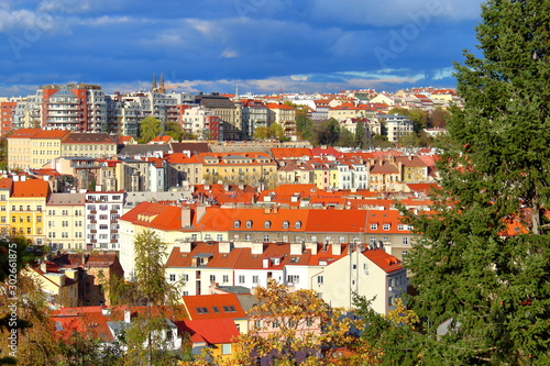 Panorama Of Prague. Houses with orange roofs, green firs with cones, yellowed trees against a blue-purple sky with heavy clouds and sunlight.