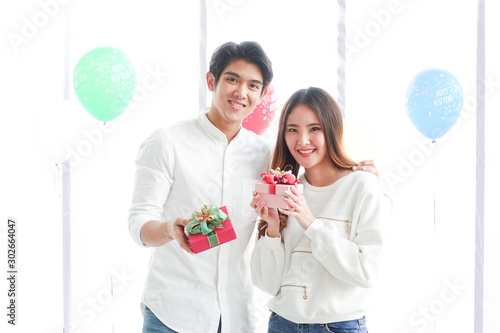 A smiling aisan couple handing out a box of present. The white room fills with colorful balloon with happt new year text. photo