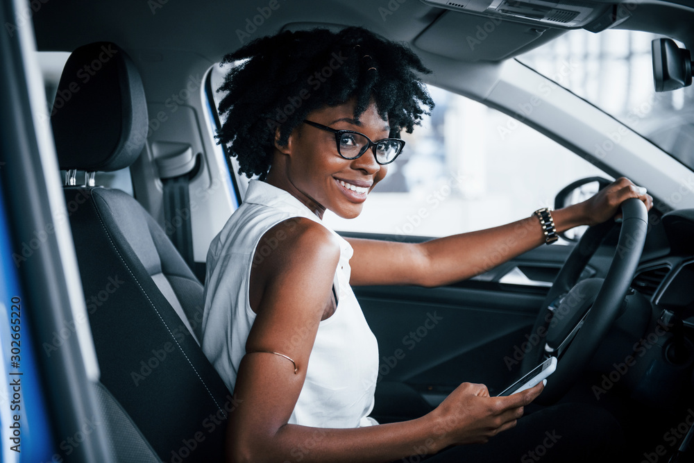 Using phone. Young african american woman sits inside of new modern car - obrazy, fototapety, plakaty 