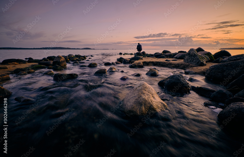 Obraz premium A person squats and looks out over the water by the sea with sunset in the background and rocks and a river in the foreground