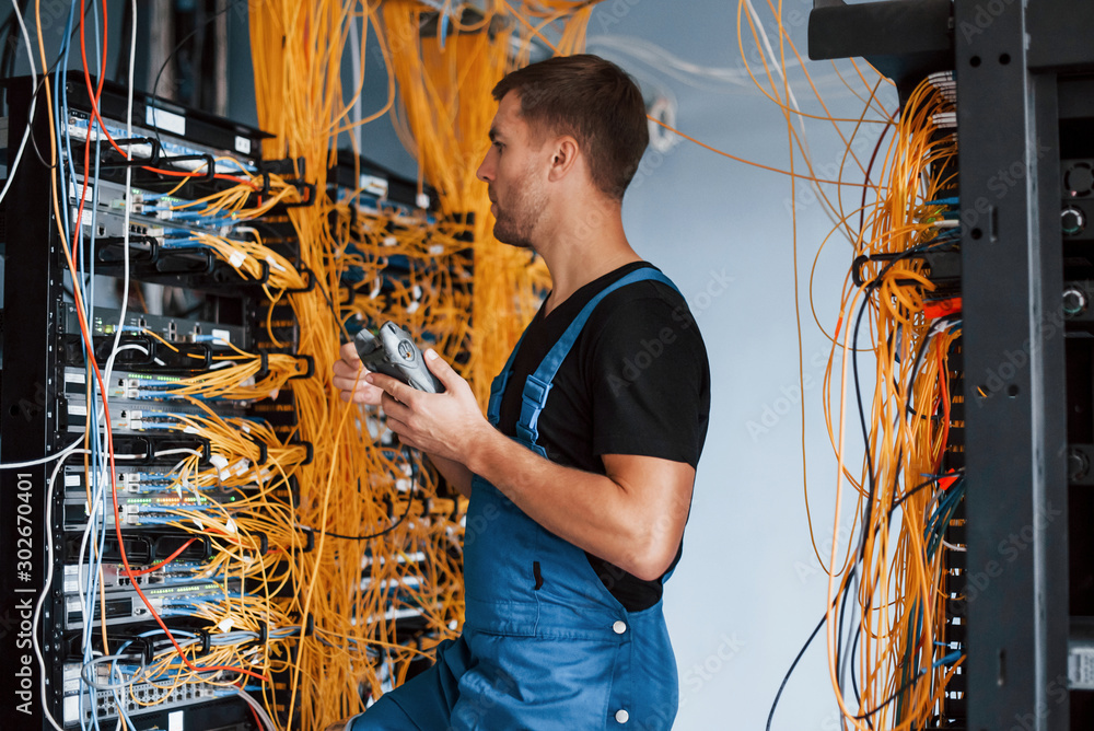 Young man in uniform with measuring device works with internet equipment and wires in server room - obrazy, fototapety, plakaty 