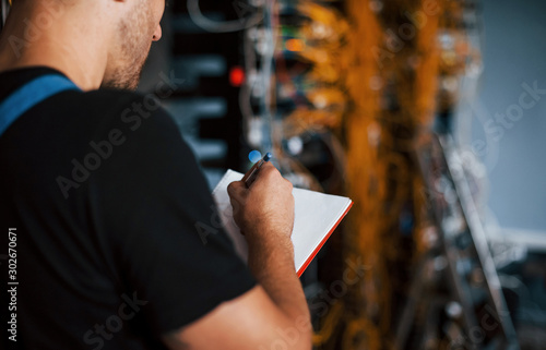 Young man in uniform with notepad in hands works with internet equipment and wires in server room