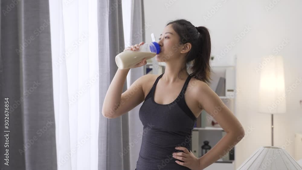 asian chinese woman in healthy lifestyle wearing sport clothes drinking bottle of milk at home by window after indoors workout. relax strong lady enjoy protein shake beverage in house living room. asian chinese woman in healthy lifestyle wearing sport clothes drinking bottle of milk at home by window after indoors workout. relax strong lady enjoy protein shake beverage in house living room.
