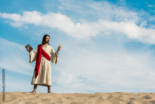 jesus holding holy bible and cross against blue sky with clouds in desert
