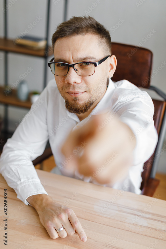 Portrait of young man sitting at his desk in the office and shows a finger at the camera