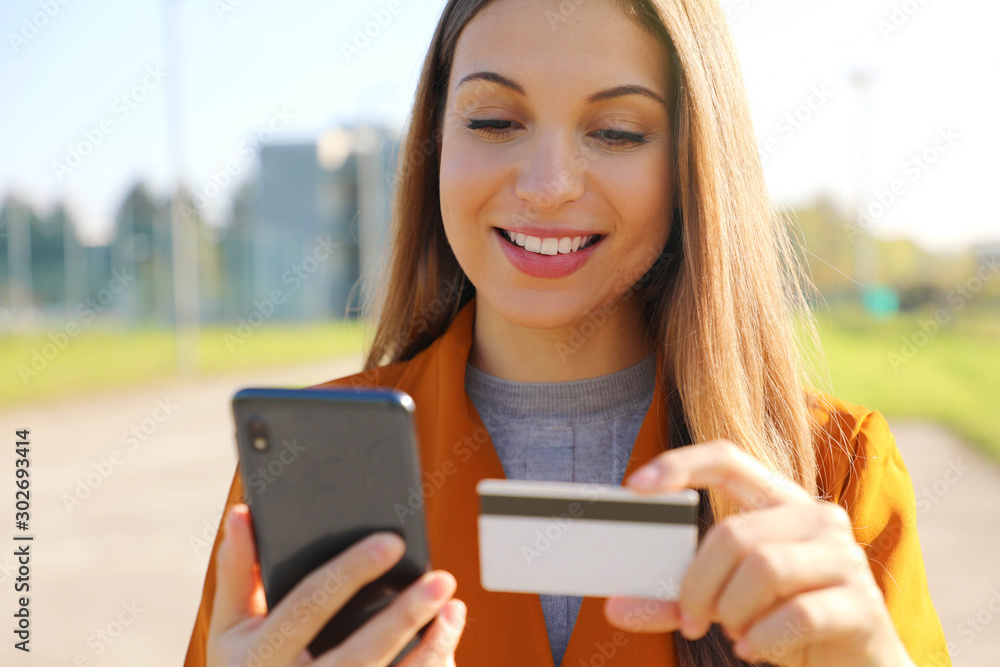 Close up of beautiful woman reading credit card number doing online payment on her smart phone outside on a sunny autumn day.