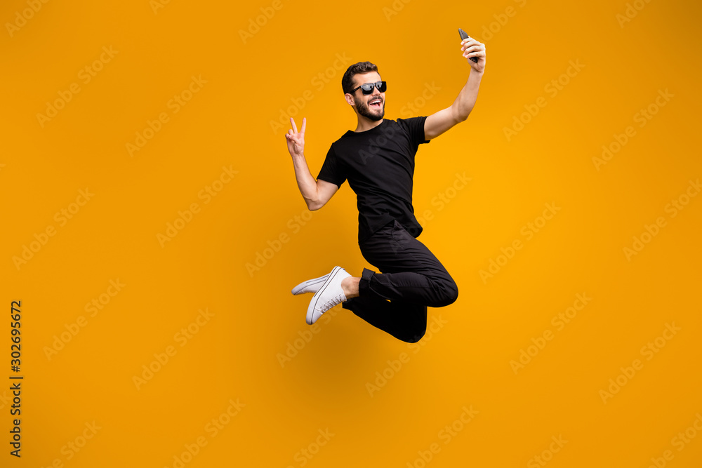 Full body photo of crazy hipster guy jumping high holding telephone making selfies showing v-sign symbol wear sun specs black t-shirt pants isolated yellow color background