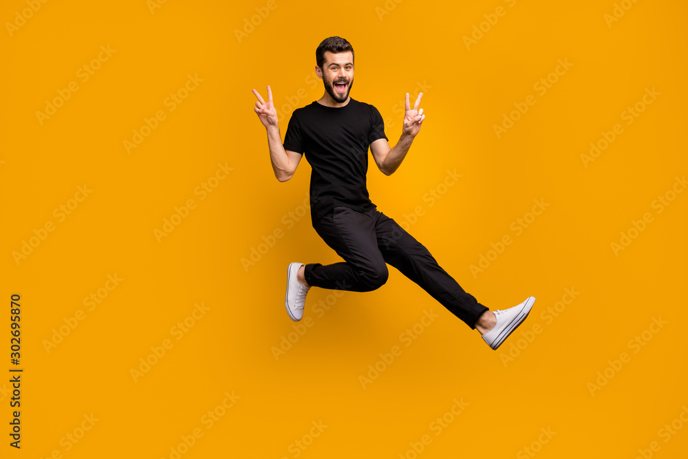 Fototapeta premium Pełne zdjęcie przystojnego podekscytowanego faceta skaczącego wysoko, podnosząc ręce, pokazującego symbole znaku v, witających przyjaciół, spotykających się na ulicy, czarna koszulka spodnie na białym tle żółty kolor tła