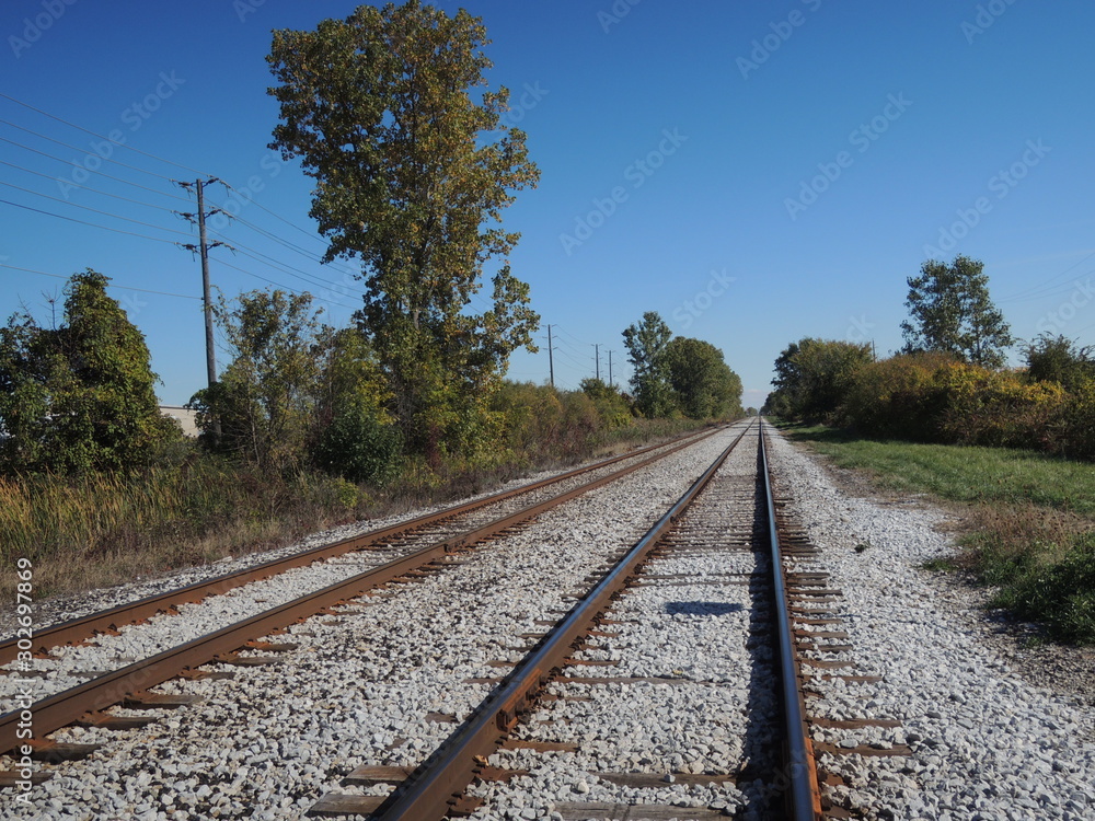 Obraz premium Rusty railway tracks in remote area. Countryside public transportation for commuters. Trees & blue sky background. Symbol for journey, new beginnings, trip, rural vacation, eco-friendly transportation