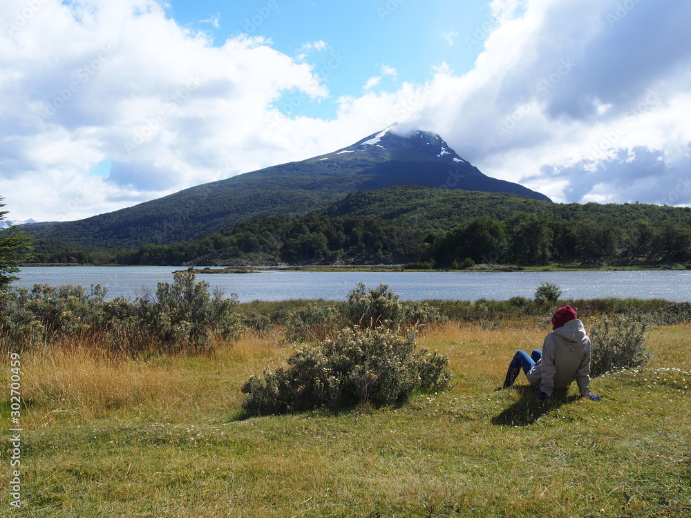 A woman sitting on a green field and gazing at the majestic mountains on a sunny day, Senda Costera (Costera Trail), Tierra del Fuego National Park, Ushuaia, Argentina A woman sitting on a green field and gazing at the majestic mountains on a sunny day, Senda Costera (Costera Trail), Tierra del Fuego National Park, Ushuaia, Argentina