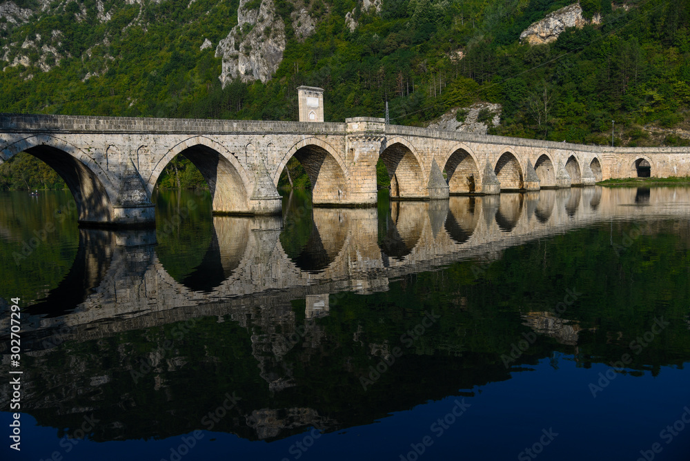 Fototapeta premium The Ottoman Mehmed Pasa Sokolovic Bridge in Visegrad, Bosnian mountains, with fantastic sky scape and river reflection. Bosnia and Herzegovina.