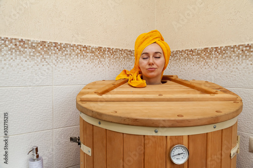 A young beautiful girl of Caucasian appearance in a cedar barrel sauna during heating and health improvement procedures with a yellow towel on her head. Health and cosmetology. photo