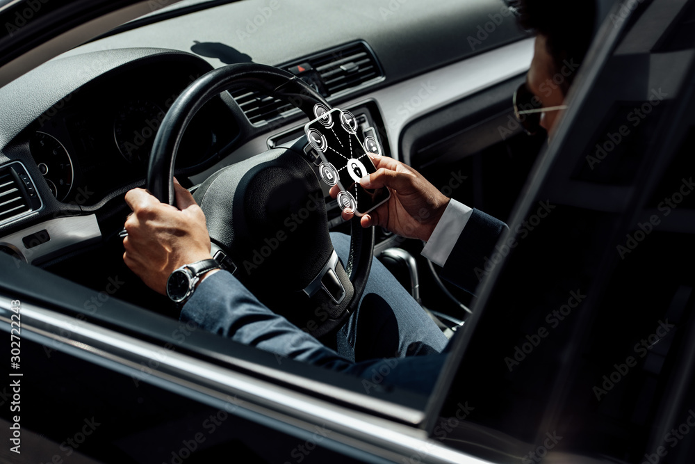 african american businessman in suit and sunglasses driving car and using smartphone with internet security icons