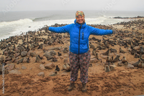 Young Caucasian red-haired happy girl traveler backpacker in a blue down jacket with spread out arms against the background of a huge colony of fur seals in Cape Kros in Namibia, Africa. photo