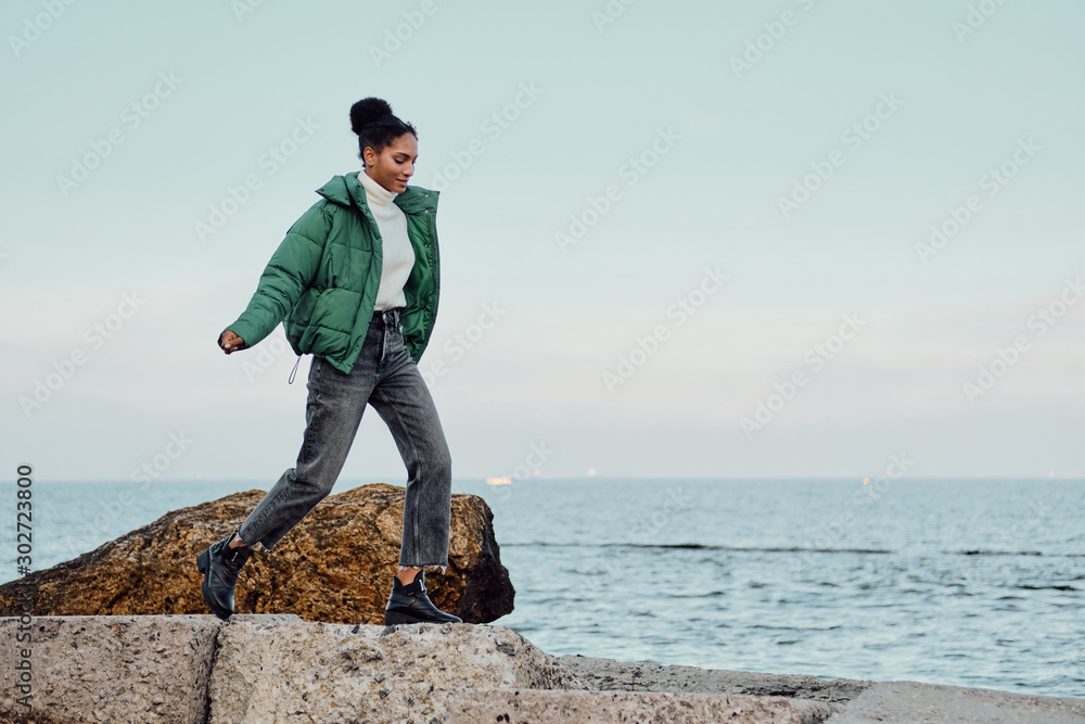 Beautiful casual African American girl in down jacket dreamily walking on stones by the sea