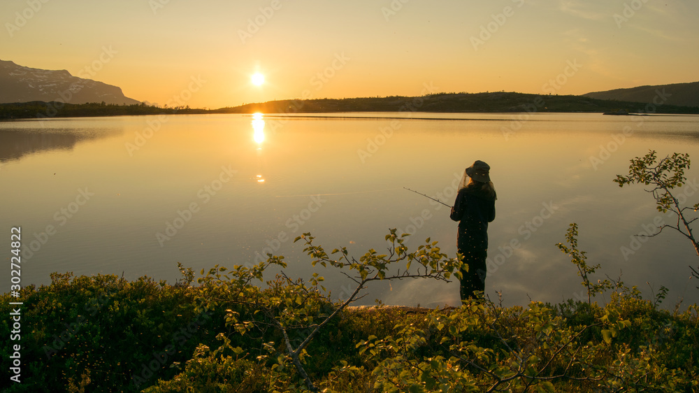 Midnightsun in Scandinavia. A girl is fishing in front of the great scenery. A mosquito hat is a necessity.