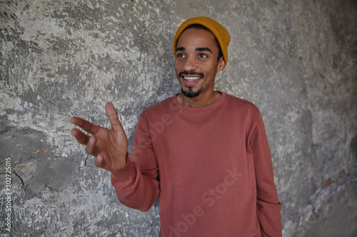 Indoor shot of joyful lovely bearded dark skinned man smiling sincerely with raised hand, looking aside while posing over concrete background, showing his pleasant emotions