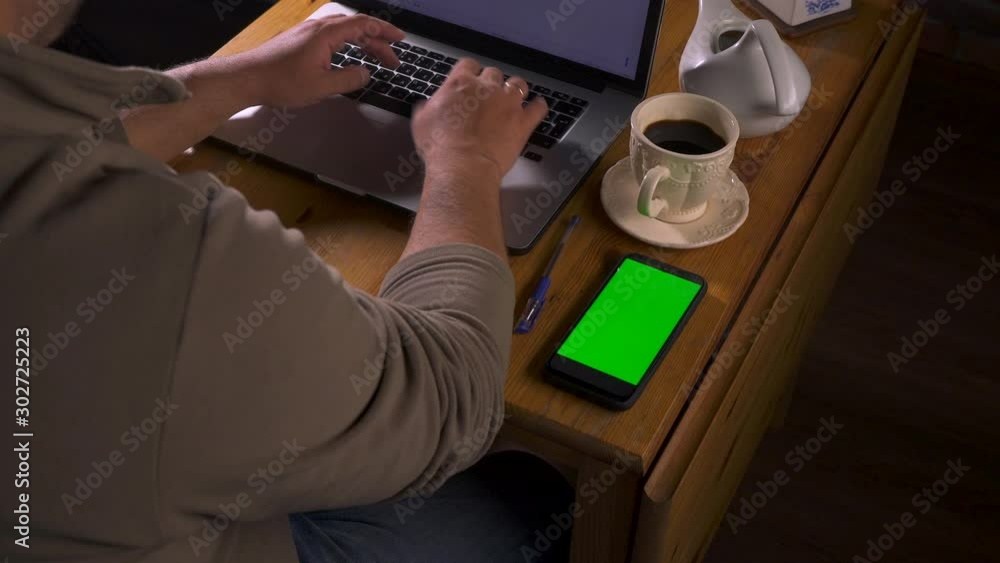 Mock-up with green screen for tracking close-up. Adult caucasian man sits at a wooden table and types text on a laptop. Nearby a phone with touch screen. Cellphone and device concept. Writer at work. Mock-up with green screen for tracking close-up. Adult caucasian man sits at a wooden table and types text on a laptop. Nearby a phone with touch screen. Cellphone and device concept. Writer at work.