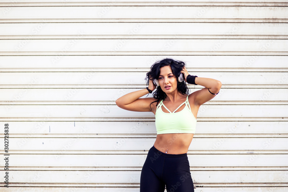 Fitness girl with long dark hair standing in front of door with headphones over her head Fitness girl with long dark hair standing in front of door with headphones over her head