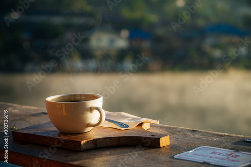 A cup of hot coffee with smoke on the wooden front of the lake view with sunlight at Ban Rak Thai,Mae Hong Son,Thailand.