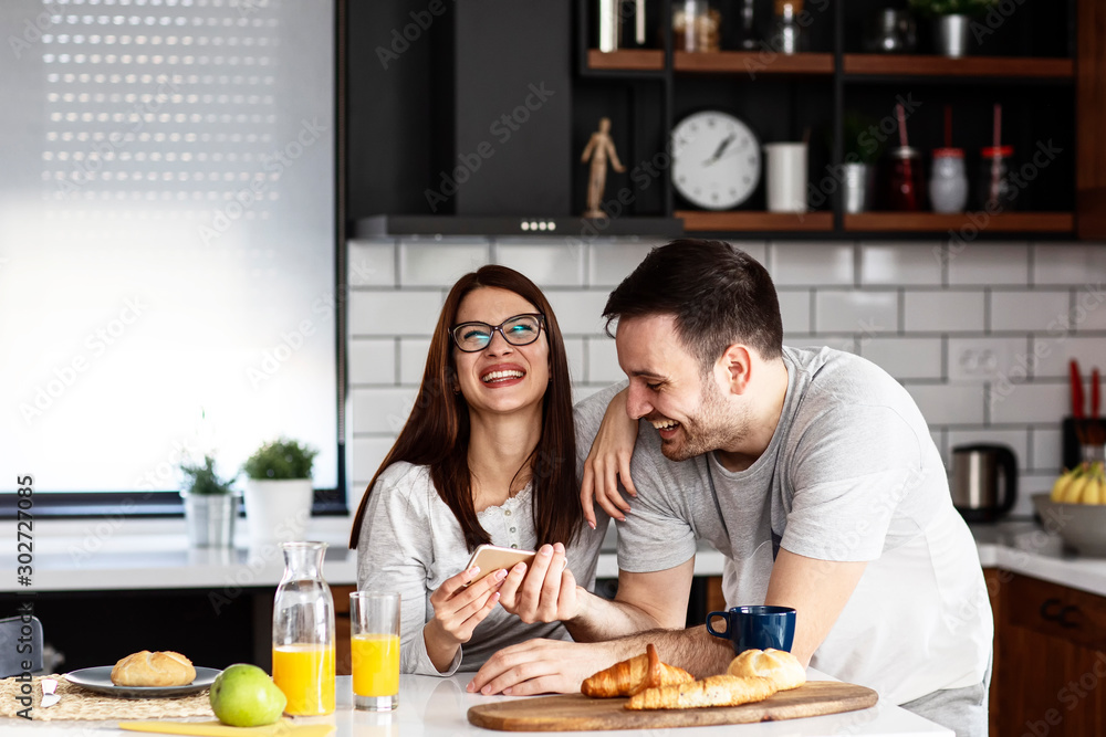 Couple in the kitchen eat breakfast with orange juice and pastry at table with mobile phone on hand smile