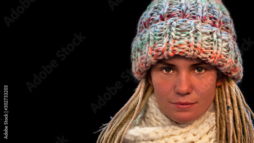 Close-up face of of green-eyed beautiful young girl with dreadlocks wearing warm winter knit hat and scarf, black isolated