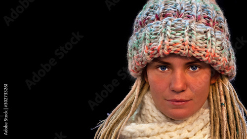 Close up face of blue-eyed beautiful young girl with dreadlocks wearing warm winter knit hat and scarf, black isolated