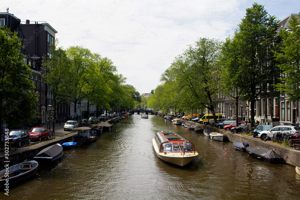 View of a cruise canal tour boat, trees, parked cars and boats, historical and traditional buildings showing Dutch architectural style in Amsterdam. It is a sunny summer day.