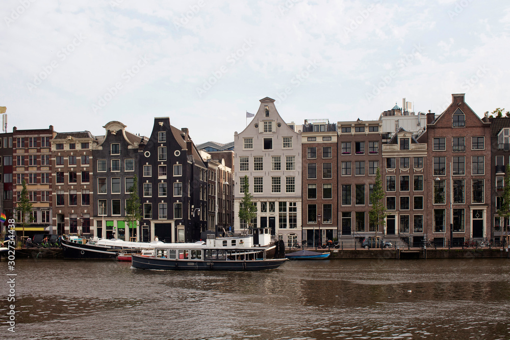 View of a boat passing on Amstel river, parked boats and historical, traditional and typical buildings reflecting Dutch architectural style in Amsterdam. It is a summer day with cloudy sky.