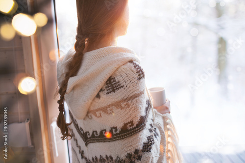 Thoughtful young brunette woman wearing nordic print poncho looking through the window, blurry winter woods snow landscape outside
